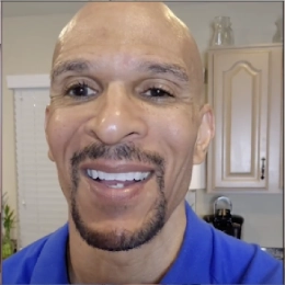 Smiling man with a trimmed beard wearing a blue polo shirt, standing in a well-lit kitchen with light-colored cabinets.
